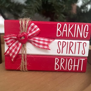 May include: A red and white wooden sign with the words "Baking Spirits Bright" in white lettering. The sign is decorated with a red and white gingham bow and a red heart button.