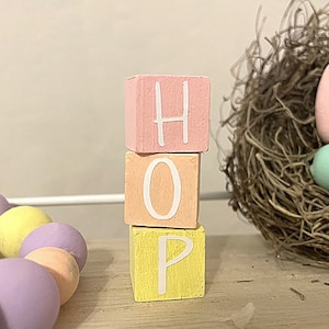 May include: Three wooden blocks stacked on top of each other, spelling out the word "HOP". The blocks are painted in pastel colors: pink, peach, and yellow. The letters are white.