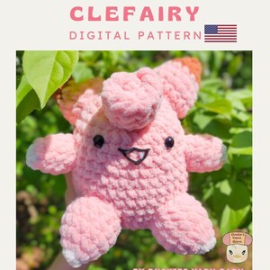 Könnte beinhalten: Rosa gehäkeltes Clefairy Plüschtier mit schwarzen Augen und einem lächelnden Mund. Das Spielzeug sitzt auf einem Bett aus grünen Blättern. Der Text "CLEFAIRY DIGITAL PATTERN" befindet sich oben im Bild. Der Text "BY DUCKIES YARN BARN" befindet sich unten im Bild.