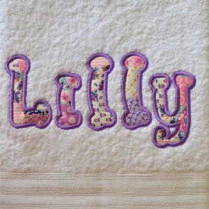 May include: White bath towel with the name "Lilly" embroidered in purple thread. The letters are filled with a patchwork of colourful fabric.