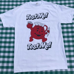 May include: A white short-sleeve t-shirt featuring a graphic of a red, round, smiling pitcher character filled with ice cubes. The character holds a smaller pitcher and a lightning bolt. The text "Test Me!" appears above and below the graphic.