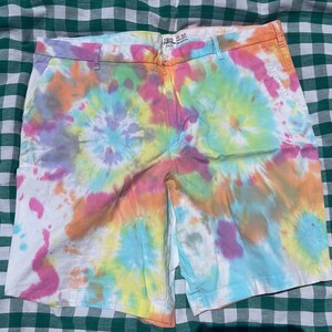 Pode incluir: Calções de comprimento médio com um padrão tie-dye vibrante em tons de rosa, laranja, amarelo, verde e azul. Os calções incluem um cós com presilhas, fecho frontal com botão e bolsos laterais. A etiqueta interior apresenta o nome Izod.