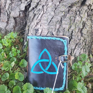 May include: A black leather journal with a blue Celtic knot design and a brown leather strap with a metal buckle. The journal is lying on a bed of green leaves.