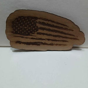 May include: A wooden plaque with a distressed American flag design. The flag features etched stars and stripes, with a natural wood grain background. The plaque has a unique, organic shape.