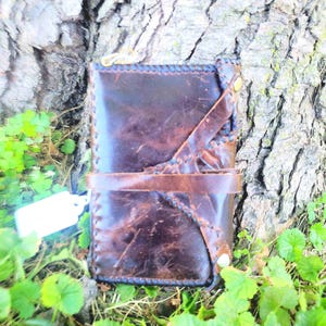 May include: A brown leather wallet with a stitched design and a metal clasp. The wallet is lying on a bed of green leaves.