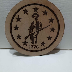 May include: A round, wooden coaster with a laser-etched design. The design features a silhouette of a colonial soldier holding a rifle, surrounded by a ring of stars. The year "1776" is etched below the soldier.