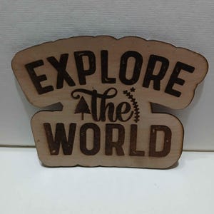 May include: Wooden sign with the words "EXPLORE the WORLD" in a dark brown font. The sign is cut in two pieces, with the top piece arched and the bottom piece rectangular. A small tree graphic is between the words "the" and "WORLD".