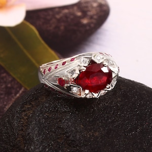 Peut inclure: Une bague en argent ornée d'un grand rubis central de forme ovale. La bague est ornée de petits rubis rouges et de diamants clairs. La monture présente des détails complexes. La bague est posée sur une pierre noire.