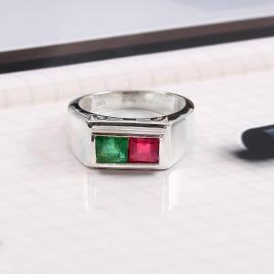 May include: A silver ring with a rectangular face. The face features two square gemstones, one green and one red. The ring is set on a white surface.
