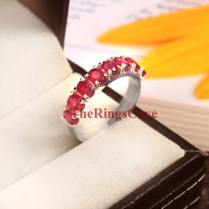 May include: A silver ring with a row of vibrant red gemstones. The gemstones are set closely together, creating a continuous band of color. The ring is displayed in a dark brown jewelry box with a white interior. The Rings Cave is printed on the ring.