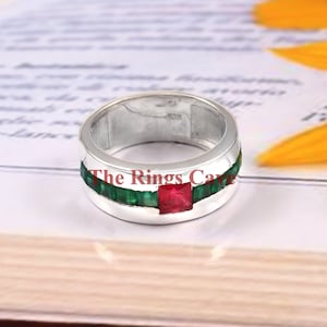 May include: A silver ring with a red square gemstone surrounded by green square gemstones. The ring is on a white surface.