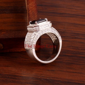 Pu&ograve; includere: Un anello d'argento con una pietra nera rettangolare al centro, circondata da pietre chiare pi&ugrave; piccole. Anche la fascia &egrave; incastonata con pietre chiare. Il testo "The Rings Cave" &egrave; visibile sull'anello.