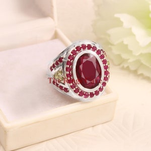 Op de afbeelding: Een zilveren ring met een grote ovale robijn, omringd door kleinere robijnen. De ring heeft een zilveren band met een decoratief ontwerp en twee kleinere groene edelstenen aan weerszijden van de robijn.
