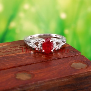 Op de afbeelding: Een zilveren ring met een rode ovale edelsteen omringd door kleinere heldere edelstenen. De ring ligt op een houten oppervlak.