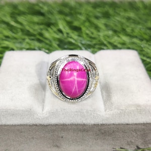 May include: A silver ring with a large, oval, pink gemstone displaying a star pattern. The ring has intricate detailing with gold accents and the text "TheRingsCafe". It sits on a white surface against a green backdrop. The ring is a statement piece.