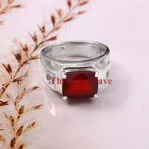 Könnte beinhalten: Ein silberner Ring mit einem großen, quadratischen, tiefroten Edelstein. Der Ring hat ein breites Band und ein geometrisches Design um den Edelstein. Der Text "The Rings Cave" ist auf dem Ring zu sehen.