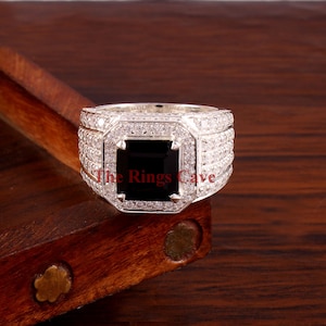 Pu&ograve; includere: Un anello d'argento con una grande pietra nera quadrata al centro. La pietra preziosa &egrave; circondata da un alone di pietre pi&ugrave; piccole e trasparenti. Anche il gambo dell'anello &egrave; incastonato con pietre trasparenti. Il testo "The Rings Cave" &egrave; visibile.