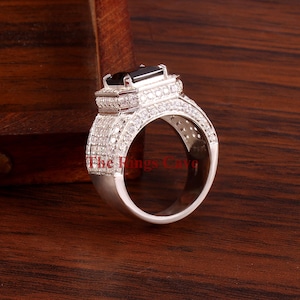 Pu&ograve; includere: Un anello d'argento con una pietra nera rettangolare al centro, circondata da pietre chiare pi&ugrave; piccole. Anche la fascia &egrave; incastonata con pietre chiare. Il testo "The Rings Cave" &egrave; visibile sull'anello.