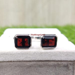 Puede incluir: Un par de gemelos rectangulares con borde plateado y centro rojo oscuro. Los gemelos se muestran sobre una superficie blanca. El texto "TheRingsCave" es visible.