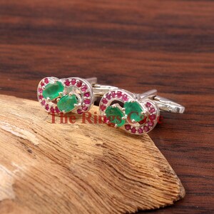 May include: Silver cufflinks with emerald and ruby gemstones. Each cufflink has two oval emeralds encircled by small rubies. The cufflinks are presented on a wooden surface.
