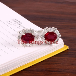 May include: A pair of silver cufflinks featuring oval-shaped red ruby gemstones encircled by a halo of smaller white gemstones.