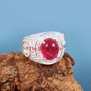Puede incluir: Un anillo de plata con una piedra preciosa roja grande de forma ovalada rodeada de piedras preciosas blancas más pequeñas. El anillo está colocado sobre una pieza de madera marrón.