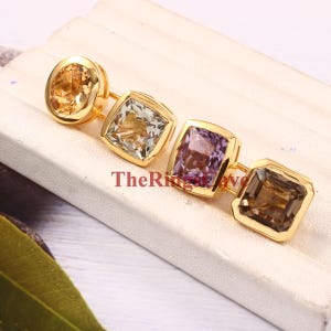 May include: Four gemstone rings with gold settings. The rings feature a round citrine, a square green amethyst, a square amethyst, and a square smoky quartz.
