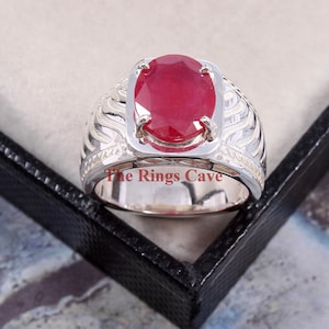 May include: A silver ring with a large, oval-shaped, red ruby gemstone set in a silver bezel. The ring band has a textured, wave-like design. The text "The Rings Cave" is visible on the ring.