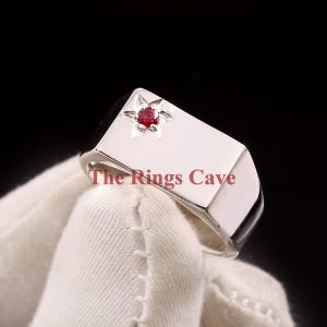 May include: A silver signet ring with a red gemstone set in a star-shaped design. The ring has a flat, rectangular top with the text "The Rings Cave" engraved on it.