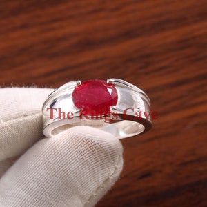 May include: A silver ring with a large, oval-shaped, red ruby gemstone. The ring has a thick band with a slightly raised edge.