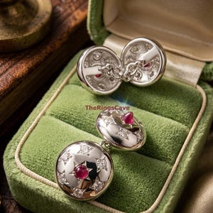 Peut inclure: Une paire de boutons de manchette argentés au design circulaire, ornés d'une pierre cabochon rouge centrale entourée de sertissages en forme d'étoile contenant des pierres claires. Le texte TheRingsCave est visible. Reliés par une chaînette.