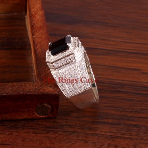 Pu&ograve; includere: Un anello d'argento con una grande pietra nera rettangolare al centro. L'anello &egrave; incastonato con piccole pietre trasparenti attorno alla pietra principale e lungo la fascia. Il testo "The Rings Cave" &egrave; visibile.