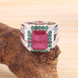 May include: A silver ring with a large, emerald-cut red gemstone surrounded by smaller green gemstones. The ring has a textured band and the words "The Rings Cave" are visible on the band.