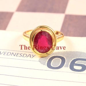 Könnte beinhalten: Ein goldener Ring mit einem großen ovalen Rubin, der in einer doppelten Zargenfassung sitzt. Der Ring liegt auf einer schwarz-weißen Schachbrettfläche.