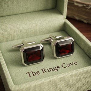 Puede incluir: Un par de gemelos plateados con piedras rectangulares facetadas de color rojo intenso. Los gemelos se presentan dentro de una caja texturizada de color verde claro con el texto The Rings Cave impreso en marrón oscuro en la base.