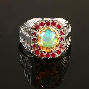 Puede incluir: Un anillo de plata con una piedra central de ópalo ovalada e iridiscente, rodeada de pequeñas gemas rojas y detalles azules. El anillo tiene un engaste dorado y detalles laterales ornamentados, reflejando la luz sobre una superficie oscura.