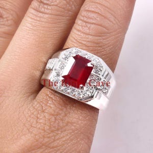 May include: A silver ring featuring a large, red, emerald-cut gemstone surrounded by smaller, clear gemstones. The ring is worn on a finger.