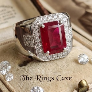 Puede incluir: Un anillo plateado con una gran gema rectangular de color rojo intenso en el centro, rodeada de gemas más pequeñas y transparentes. El anillo tiene una cara cuadrada y está sobre un fondo beige suave. El texto "The Rings Cave" es visible.