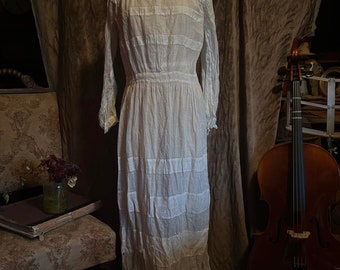 May include: A white cotton dress with a gathered skirt and long sleeves. The dress is on a dress form in front of a brown fabric backdrop.