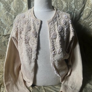 Könnte beinhalten: Ein cremefarbener Cardigan mit offener Front und aufwendigen weißen Blumenperlen und Verzierungen. Der Pullover hat einen runden Ausschnitt, lange Ärmel und gerippte Bündchen. Der Hintergrund ist ein goldfarbener Stoff mit Muster.