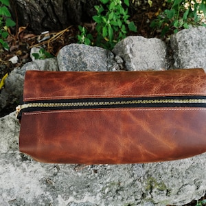 May include: A brown leather zippered pouch with a gold zipper. The pouch is sitting on a rock.