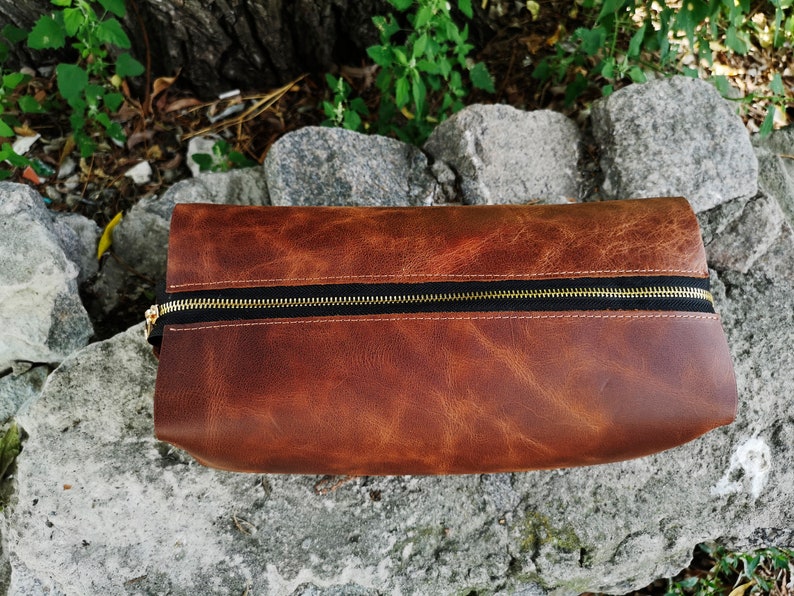 May include: A brown leather zippered pouch with a gold zipper. The pouch is sitting on a rock.