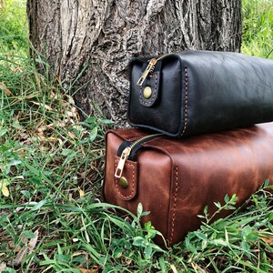 May include: Two leather toiletry bags, one brown and one black, with gold zippers and stitching. The bags are sitting on a bed of green grass.