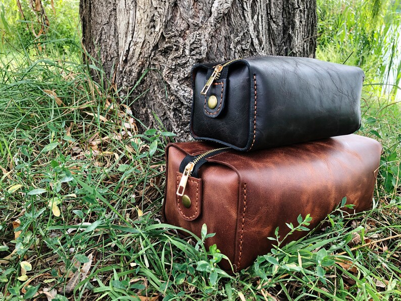 May include: Two leather toiletry bags, one brown and one black, with gold zippers and stitching. The bags are sitting on a bed of green grass.