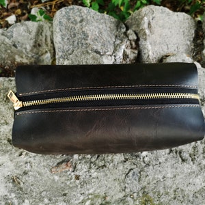 May include: A dark brown leather zippered pouch with a gold zipper. The pouch is lying on a gray, textured surface.