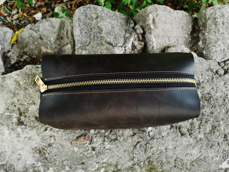 May include: A dark brown leather zippered pouch with a gold zipper. The pouch is lying on a gray, textured surface.