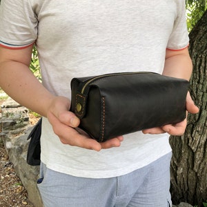 May include: A black leather travel toiletry bag with a zipper closure and orange stitching. The bag is held in the hands of a person.