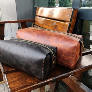 May include: Two leather zippered pouches, one brown and one black, sitting on a wooden bench.