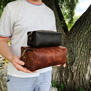 May include: Two leather travel bags, one brown and one black, with gold zippers and hardware. The bags are stacked on top of each other and being held by a person.