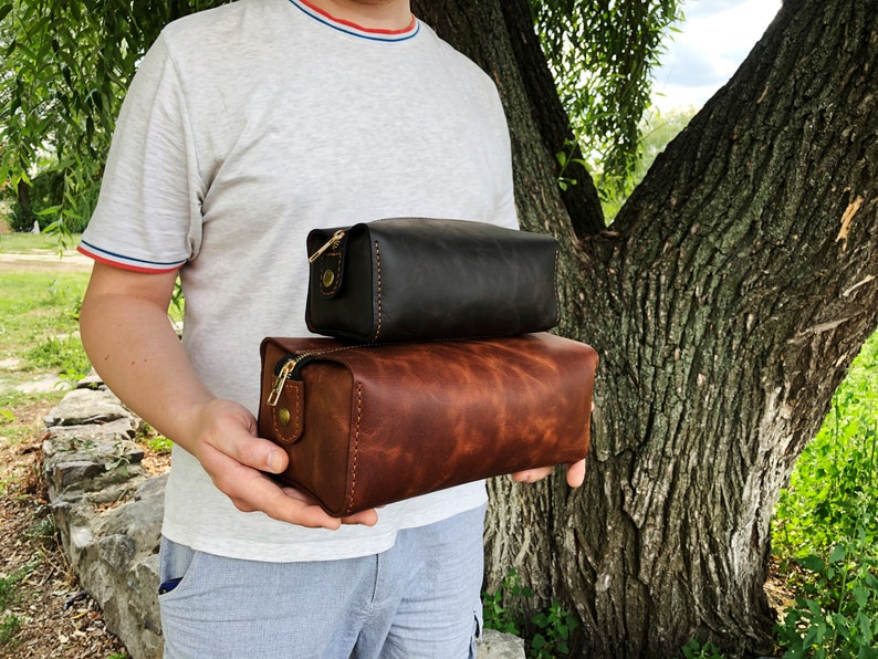 May include: Two leather travel bags, one brown and one black, with gold zippers and hardware. The bags are stacked on top of each other and being held by a person.
